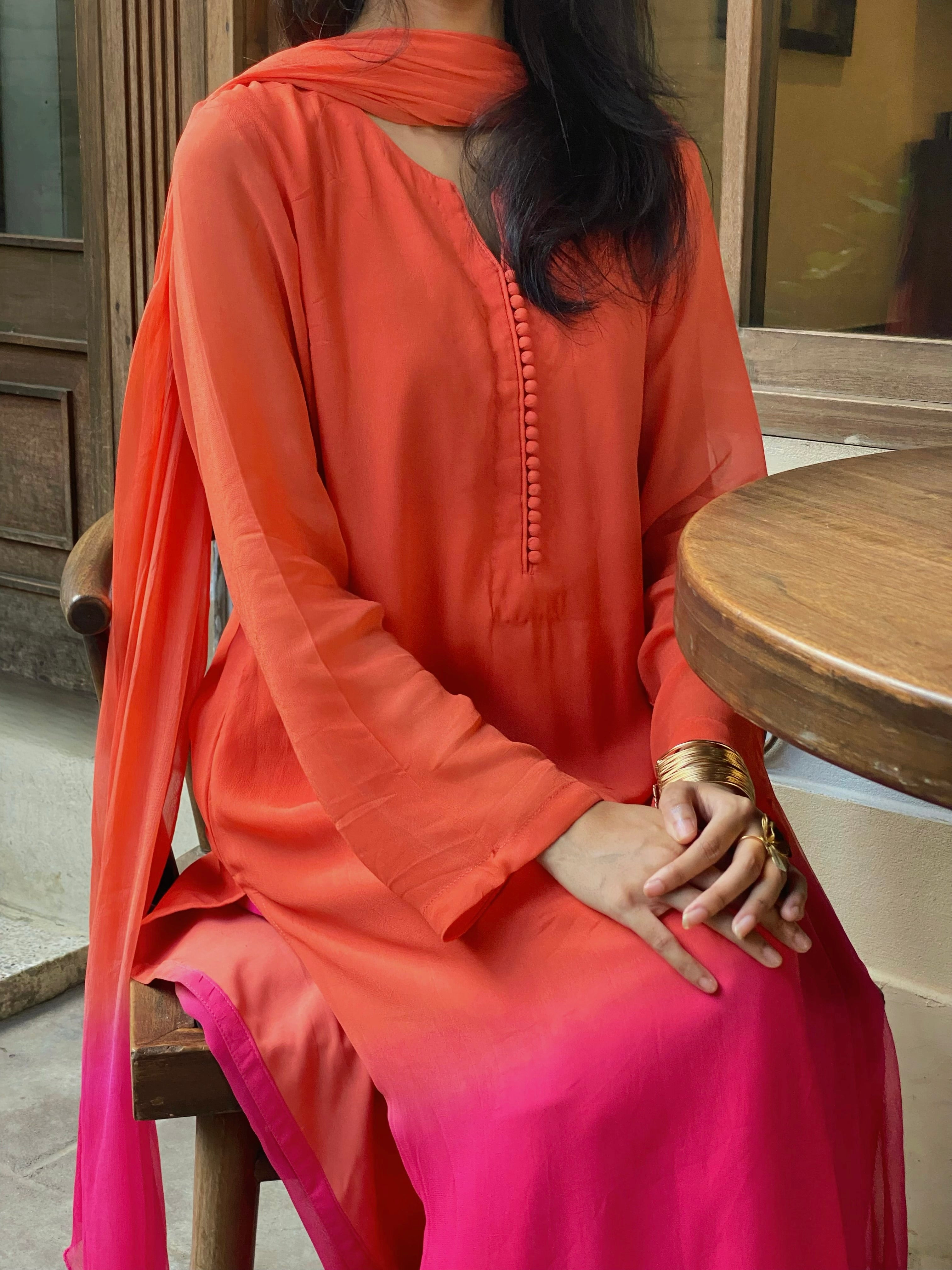 Person wearing an orange and pink traditional outfit sitting on a wooden stool.